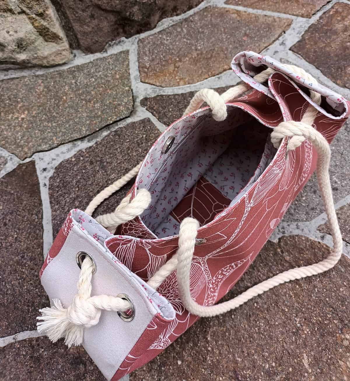 Schultertasche aus Stoff mit Seilgriffen, auf Kopfsteinpflaster liegend.